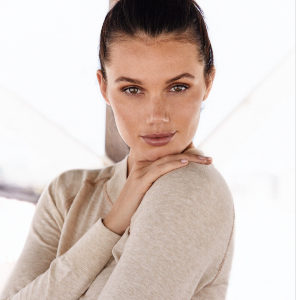 Image of a Woman  in a relaxed pose, wearing an up-do hairstyle and relaxing in her Restore Cotton Crewneck Sweater in Lynx Beige. There is an early daylight fog in the background on a beach in South Miami FL