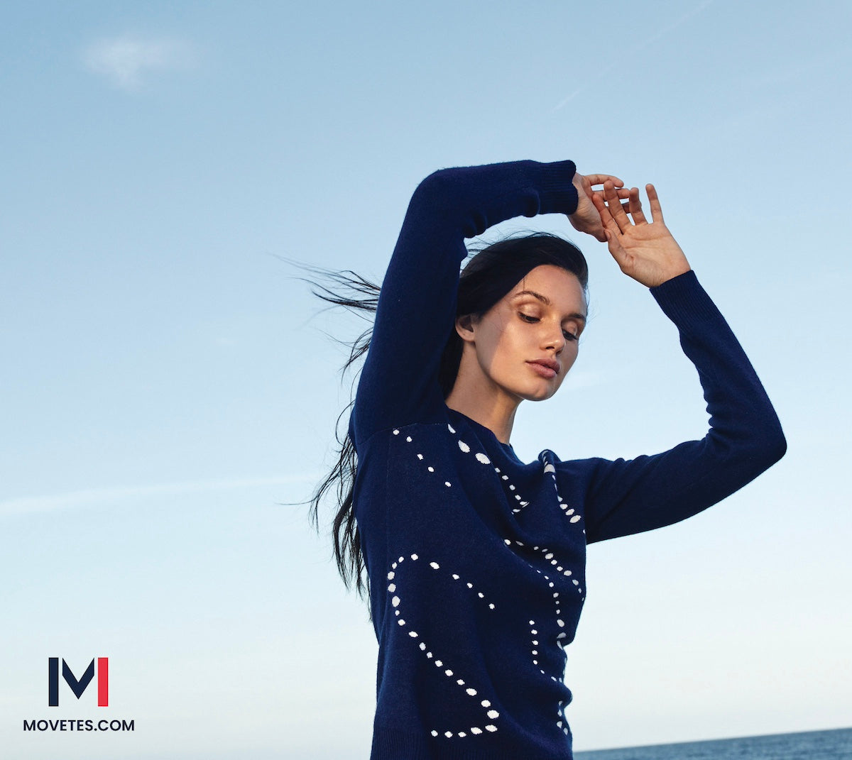 This image shows a woman in freeform, wearing the Aubrey Intarsia Varsity Crewneck Sweater,  in Admiral Navy,with a string of pearls motif. The photo was taken in South Miami Beach during a beautiful morning sunrise.