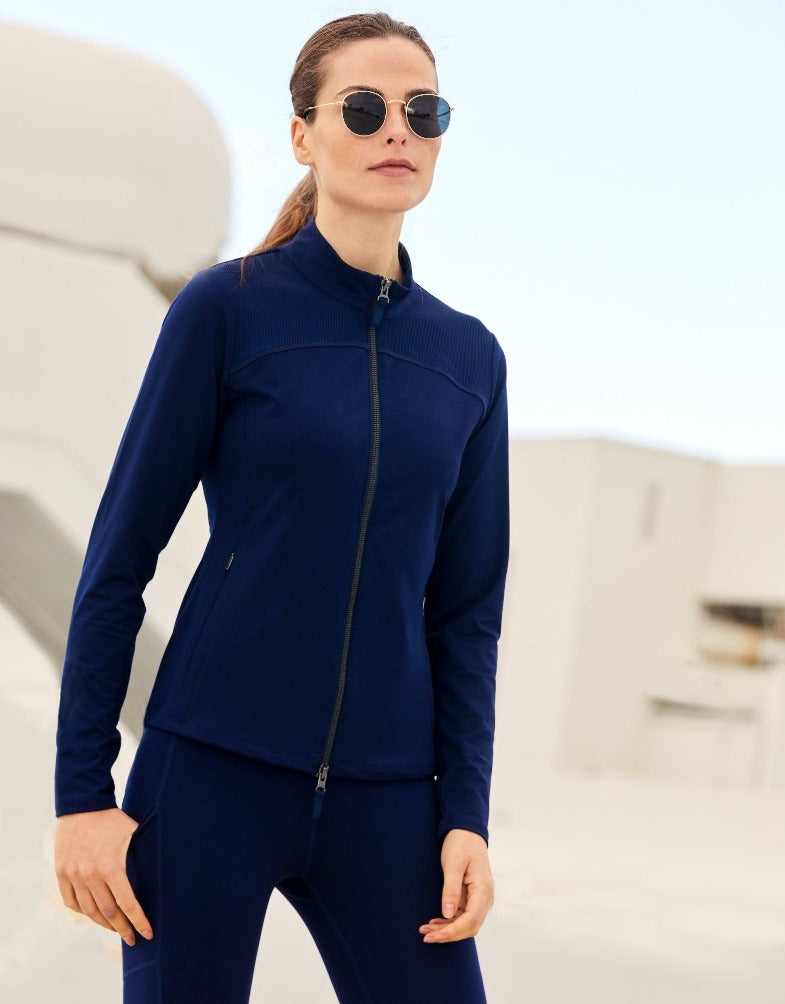 Image shows a woman wearing the Navy Jersey Powerstretch Jacket with Navy Nova Legging in an outdoor setting. Sustainable Luxury crafted using the highest quality Recycled textiles and zipper trims. Features a 2-way full front zipper and roomy side zip pockets.