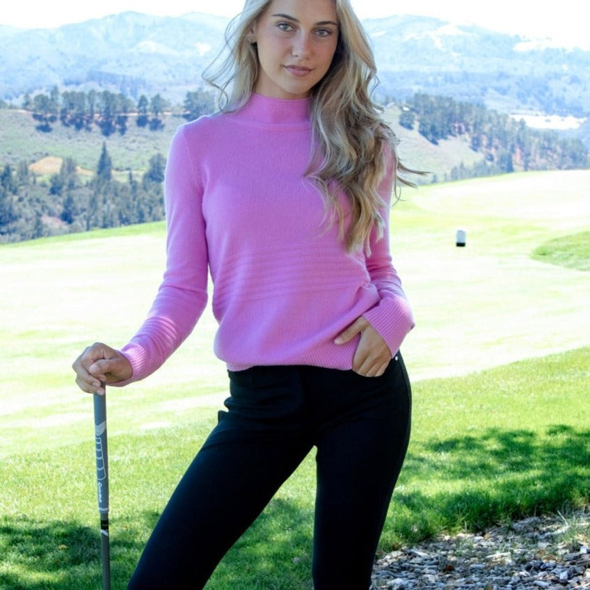 This image shows a woman wearing the Isabella Cashmere Golf Sweater in Carnation Pink, Paired with the Black PowerStretch Jersey Tailored Eliza 5-Pocket Pant. Sustainable Luxury that Moves With You.