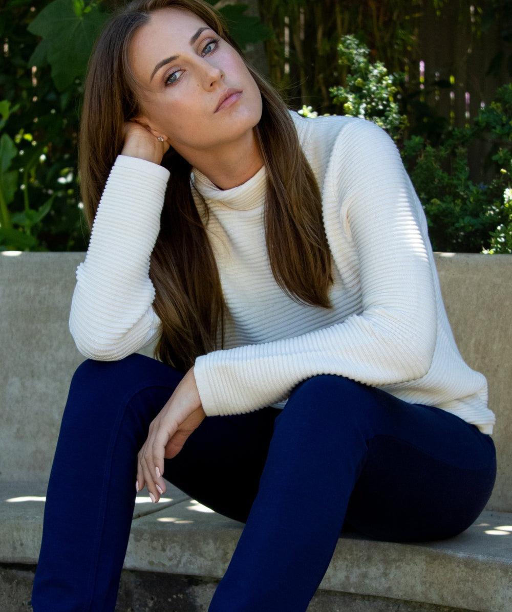 Embracing elegant versatility and quiet style with the Ellie Cotton Rib Funnel Neck Sweater. Sustainable and sumptuous; crafted of Pure Cotton. This image shows a woman wearing the Ellie Cotton Link Funnel Neck Pullover in Natural Ivory, paired with a Navy 5-Pocket Stretch Jersey Eliza Pant.